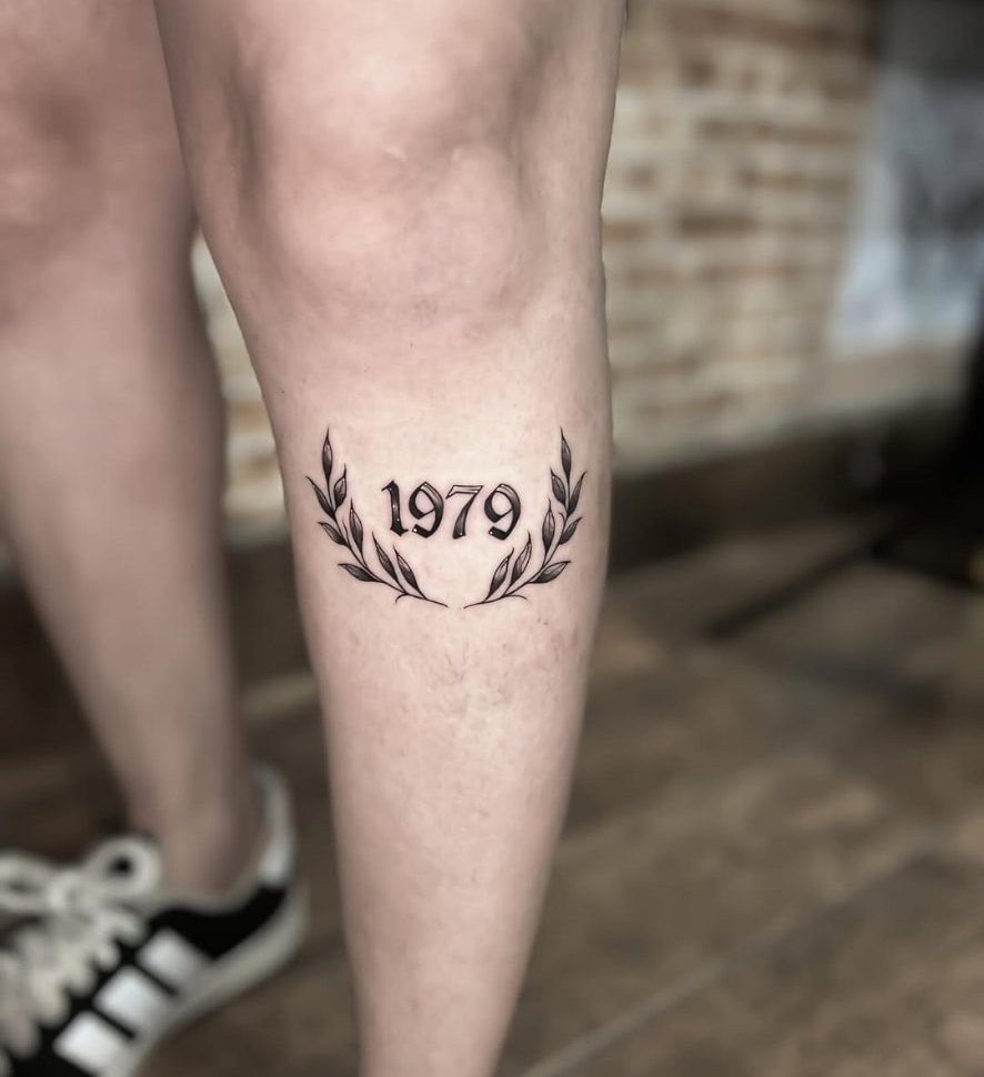 Tatuagem de data 1979 com ramos na panturrilha